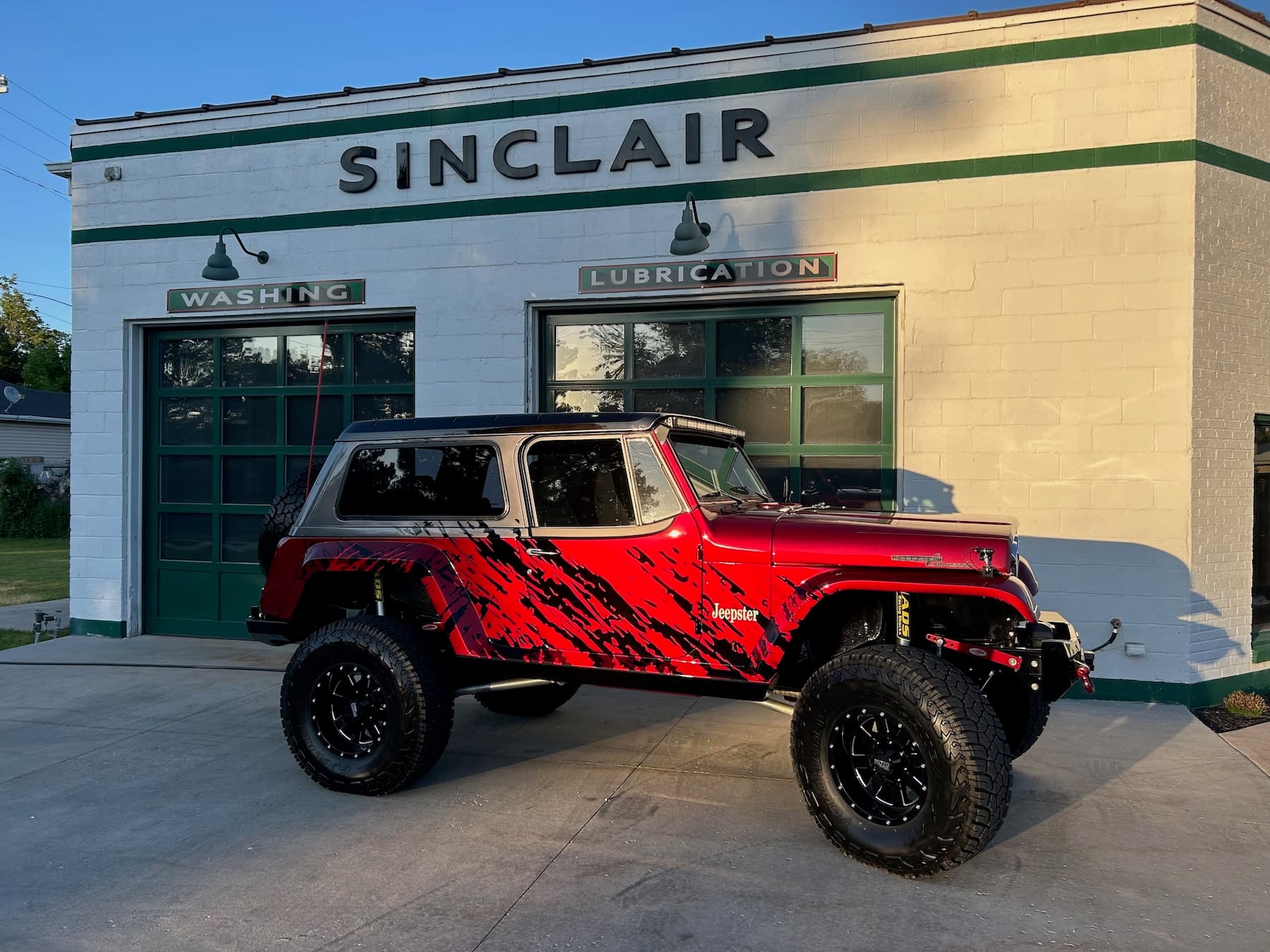 Lifted red Jeepster Commando with black graphics parked in front of a vintage Sinclair service station.