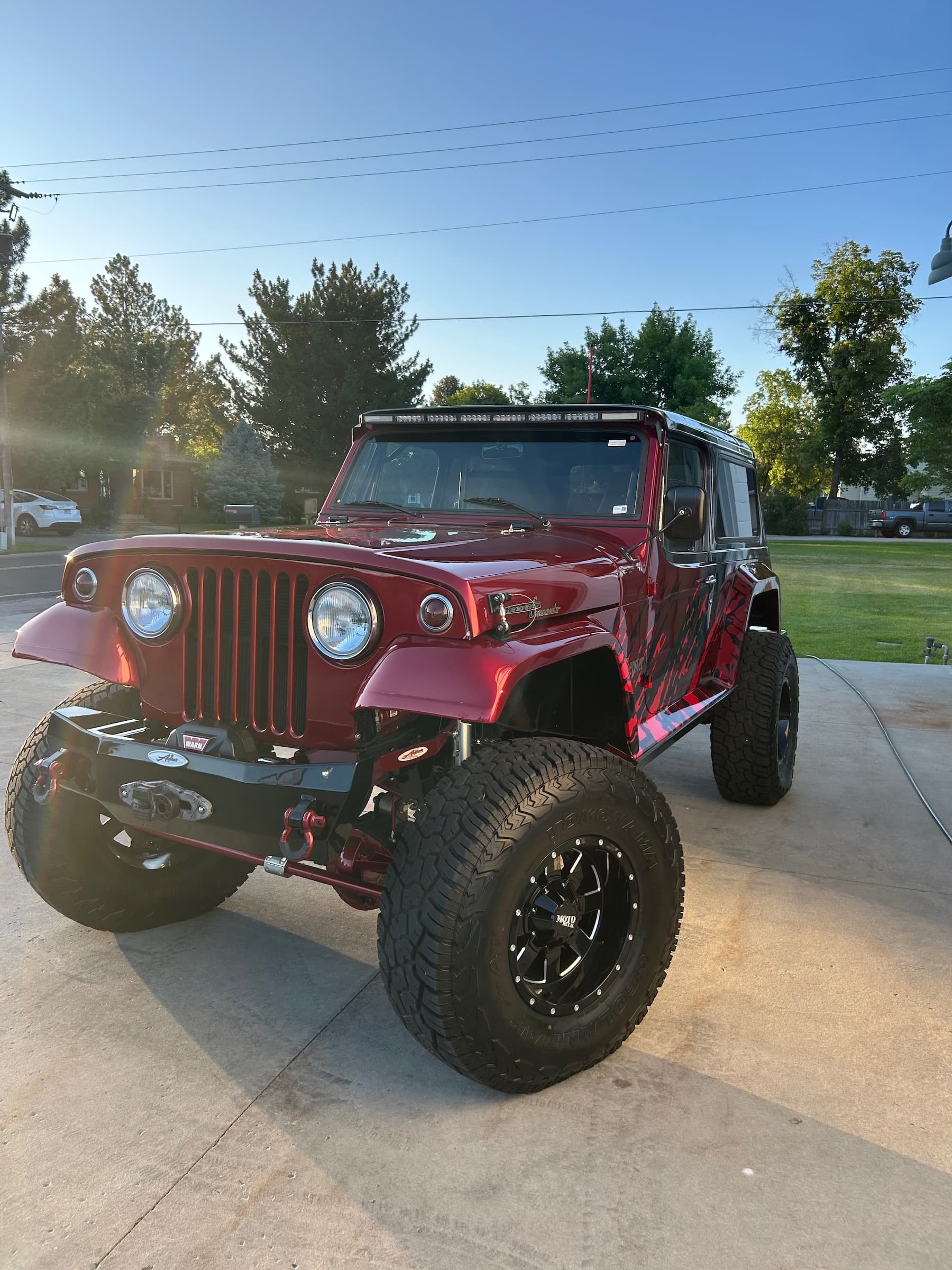 Red lifted Jeep Commando with oversized tires and black graphics on a sunny driveway.