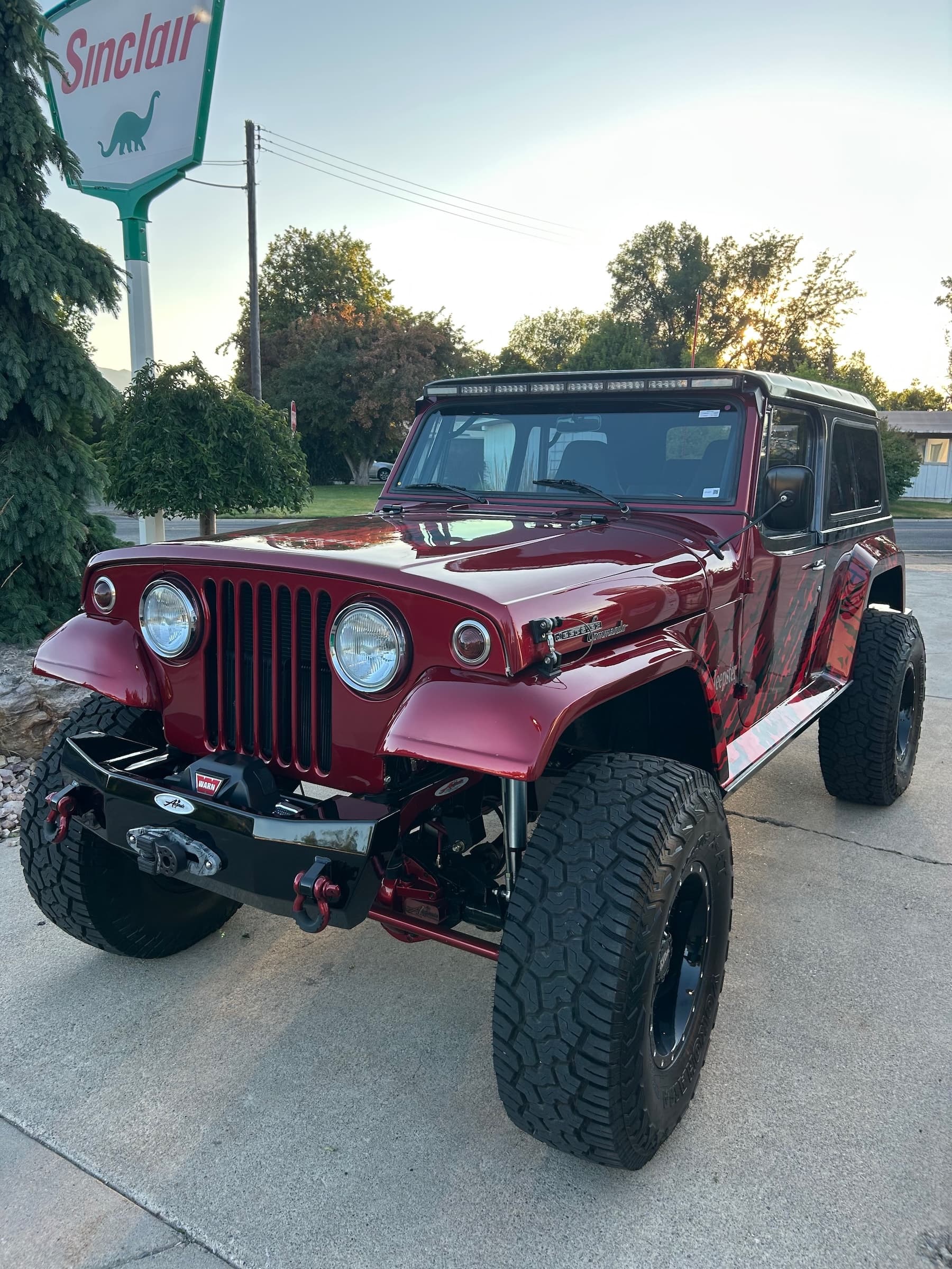 Modified red Jeep Commando with large off-road tires parked outdoors near a Sinclair sign.